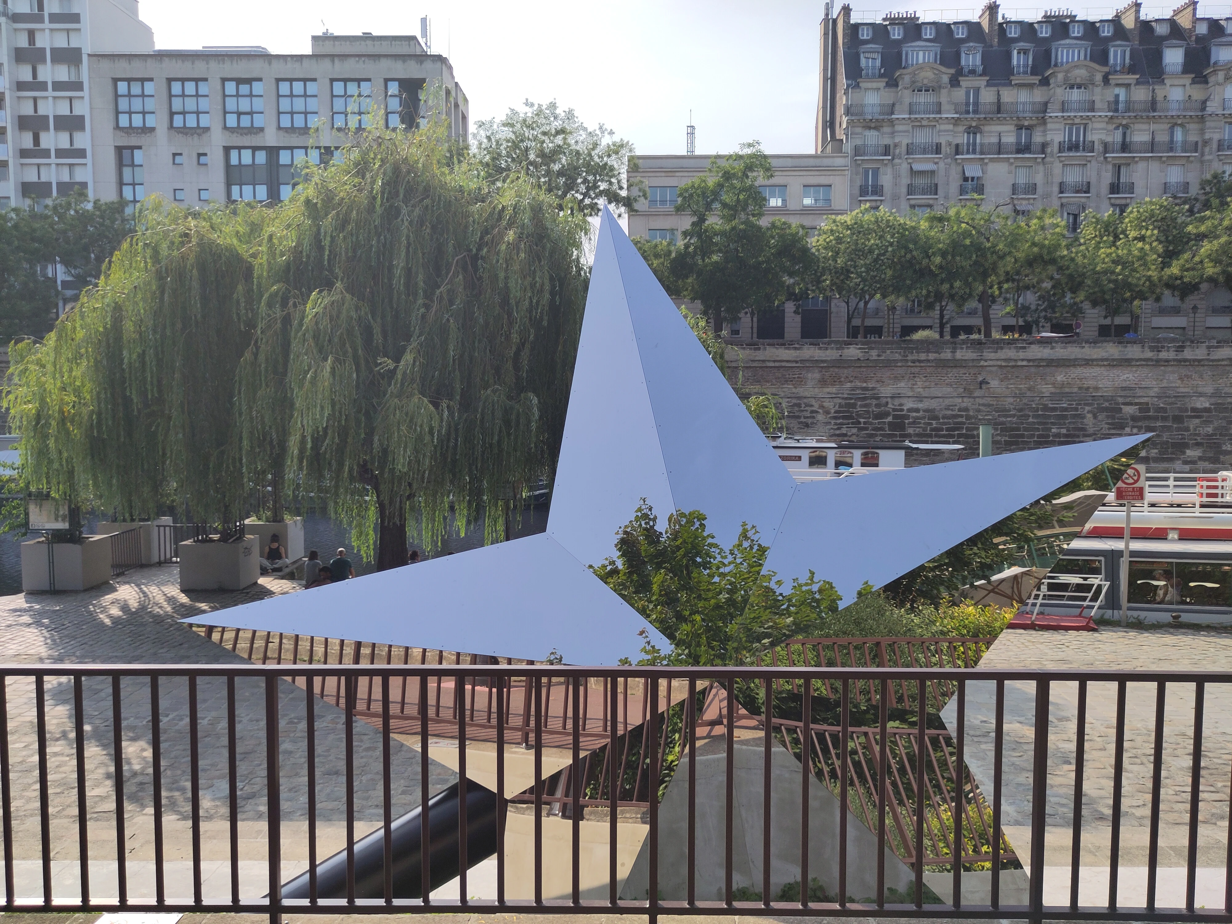 étoile face miroir en premier plan, derrière une ballustrade. on vout le quai, un bateau, un saule pleureur et les immeuble de l’autre côté du bassin.