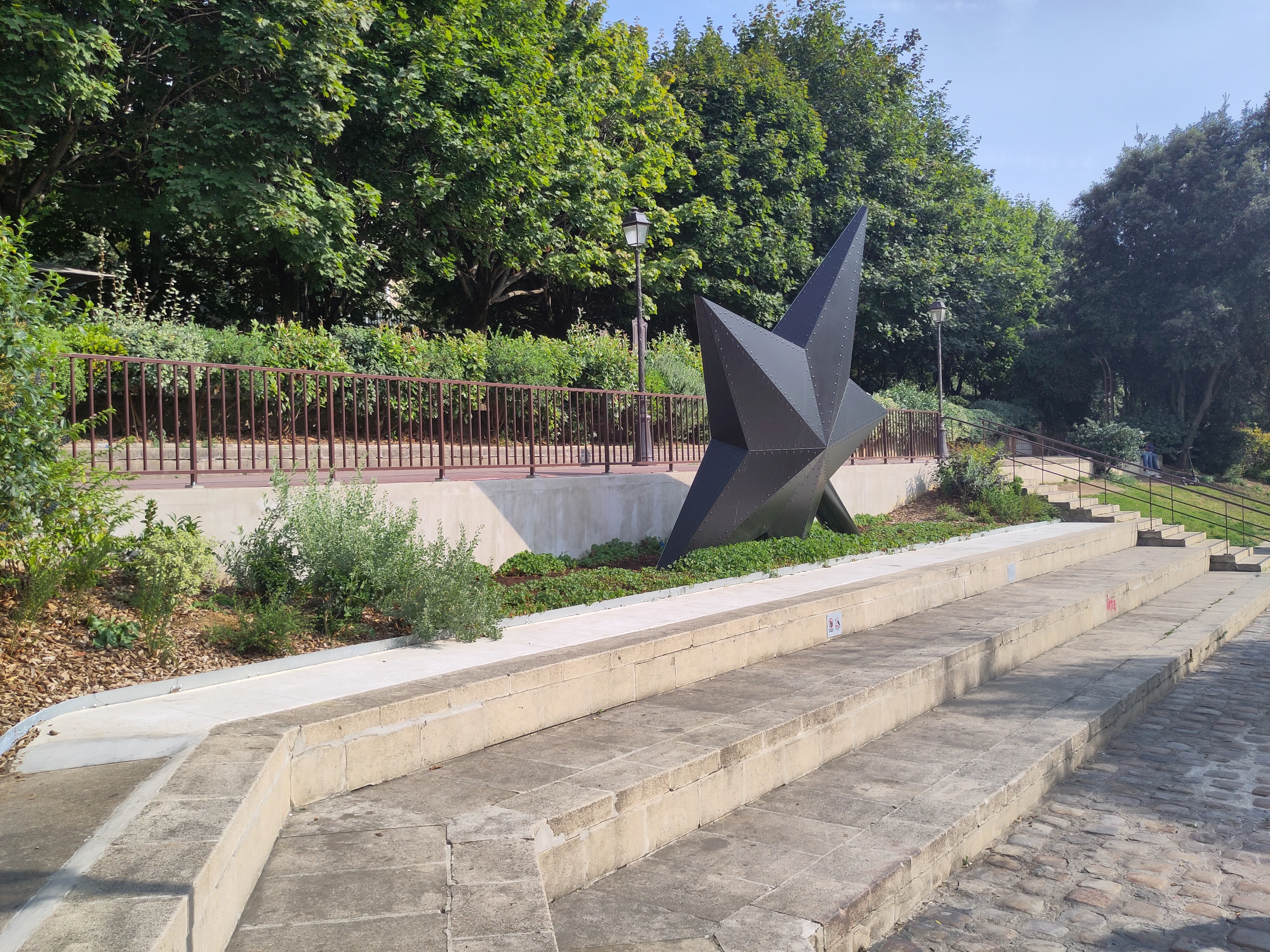 étoile vue du côté noir, en second plan. Elle est en haut de 3 marches formant un gradin de pierre en premier plan. On voit des arbres, lampadaires, ballustrades, pavé…