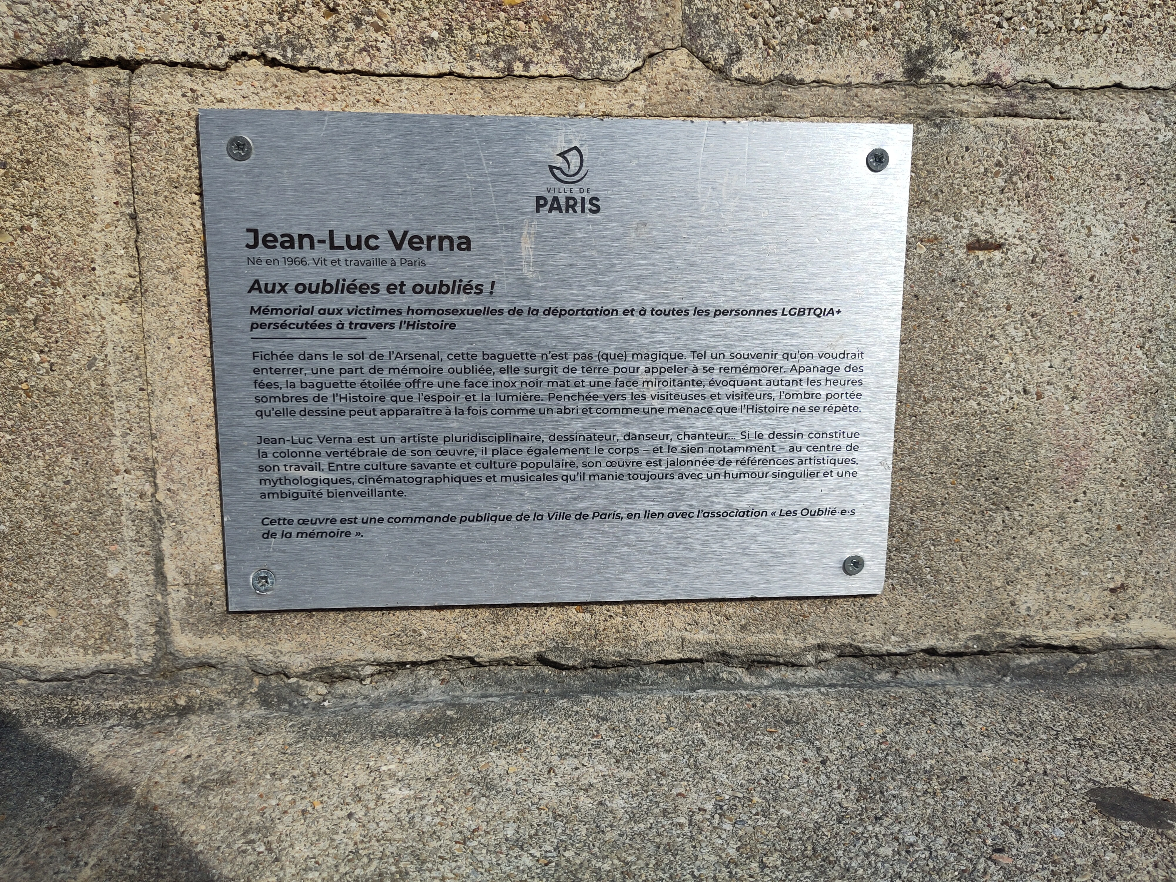 plaque métallique grise claire visée sur une pierre. Texte en noir, écrit plus bas dans la page